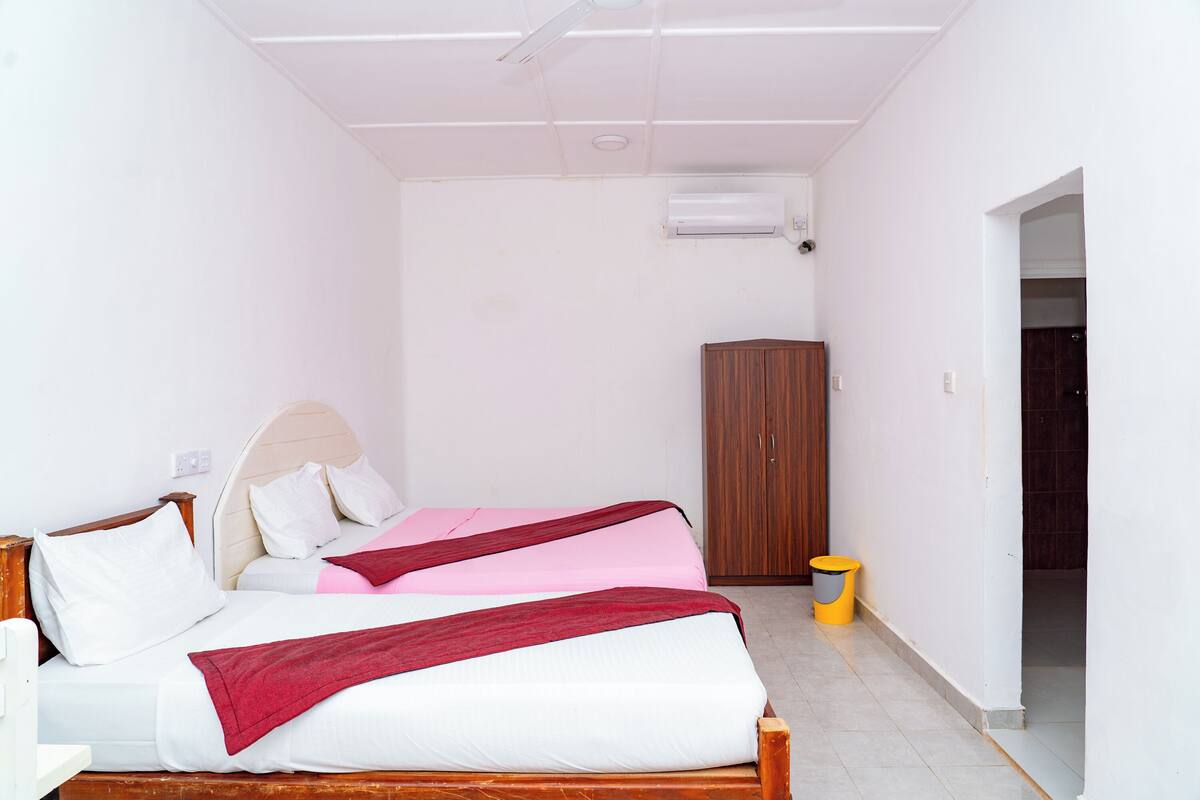 A room with two beds, a wooden wardrobe, and a trash bin.
