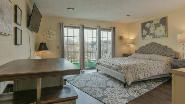 A bedroom with a bed, a desk, a chair, a lamp, and a large window with curtains.