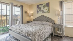 A bedroom with a large bed, two bedside tables, a window with curtains, and a wall-mounted lamp.
