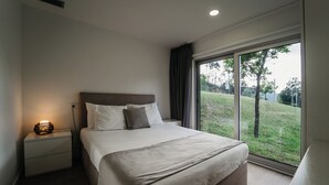 A modern bedroom with a large bed, a bedside table with a lamp, and a view of a grassy area through a large window. - Welcome Douro (Cinfães)