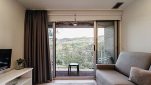 A modern living room with a sofa, a TV on a stand, a small stool, and a sliding glass door leading to a balcony with a view of trees and hills. - Welcome Douro (Cinfães)