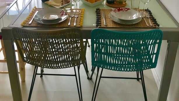 Een eettafel gedekt met borden, bestek en een fles, omringd door rieten stoelen, met uitzicht op groen buiten.