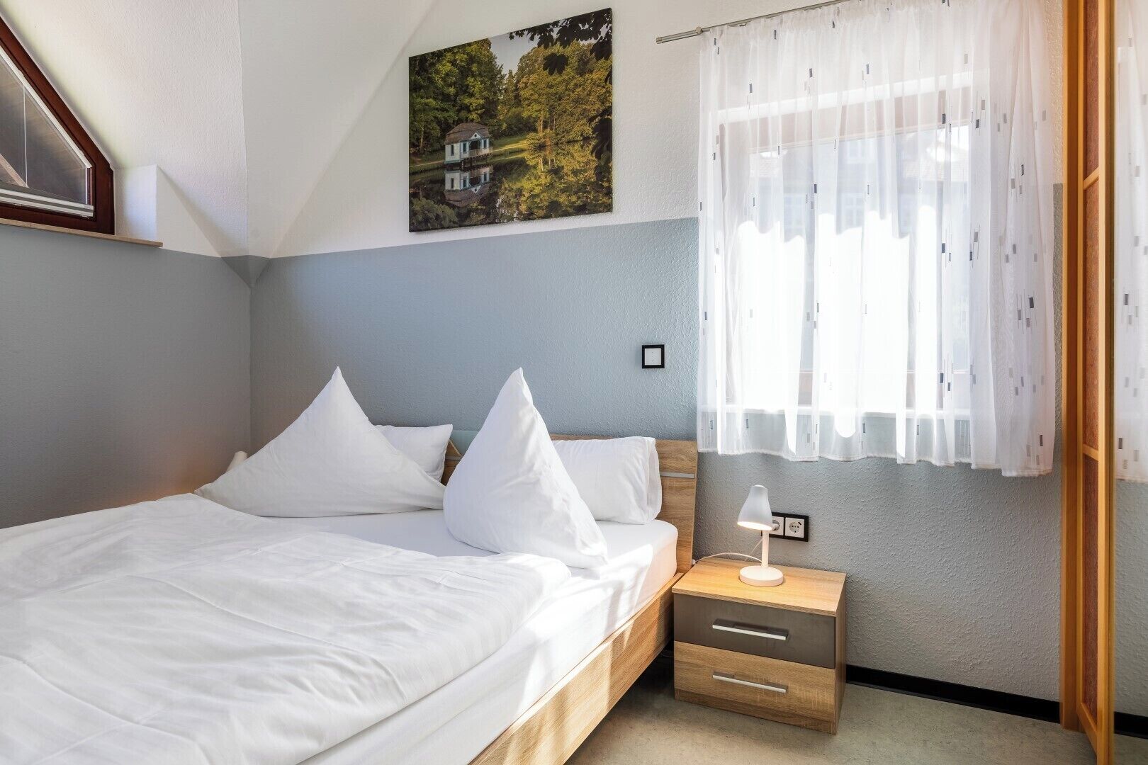 Ein Schlafzimmer mit einem Bett, Kissen, einem Nachttisch mit Lampe, einem Fenster mit durchsichtigen Vorhängen und einem Gemälde an der Wand.