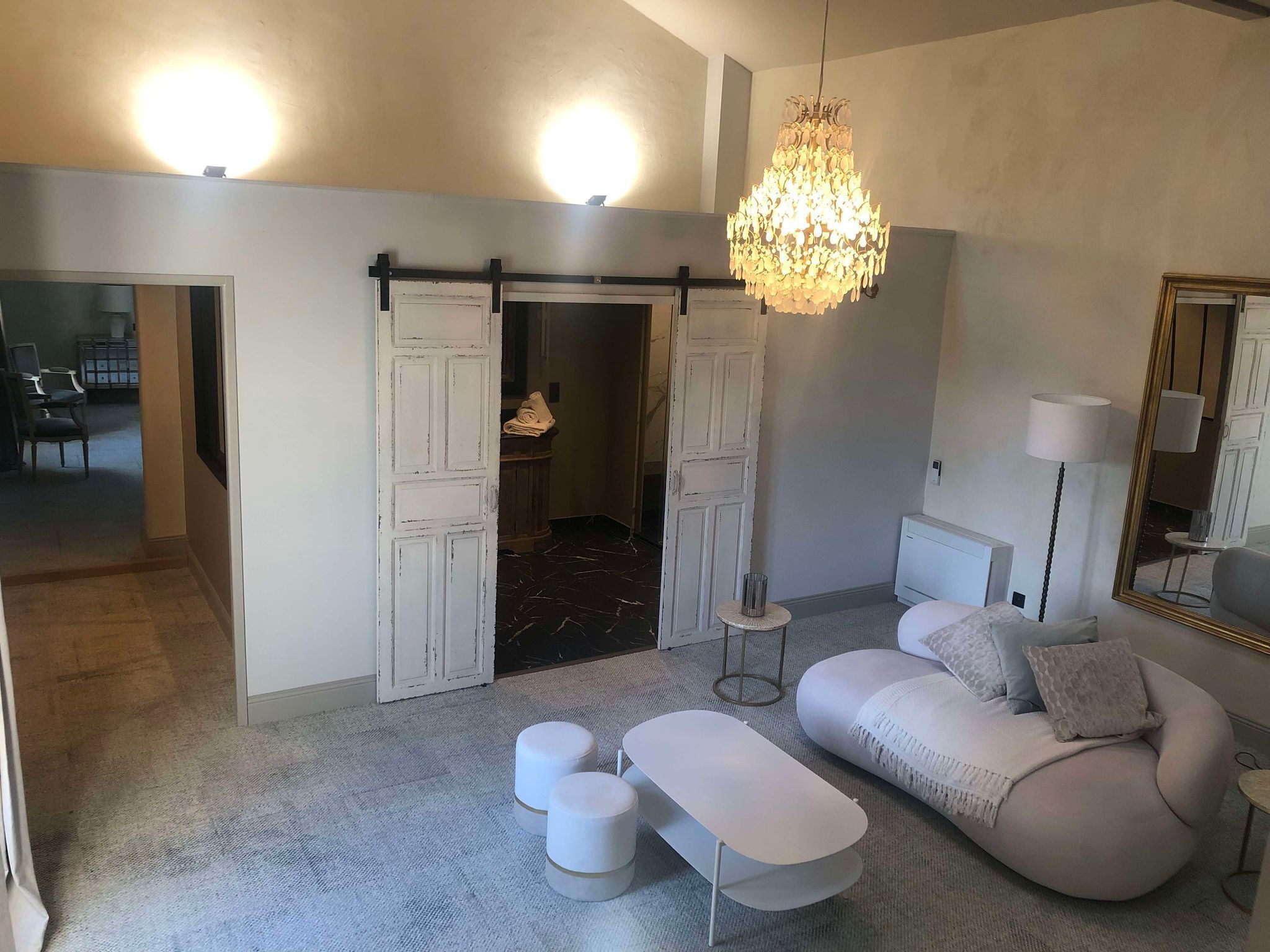 Ein modernes Wohnzimmer mit einem weißen Sofa, einem Hocker und einer Deckenleuchte. Ein großes Spiegelbild und eine Tür zu einem weiteren Raum sind ebenfalls vorhanden.