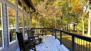 A wooden deck with two chairs and a trash can, surrounded by trees and a fence.