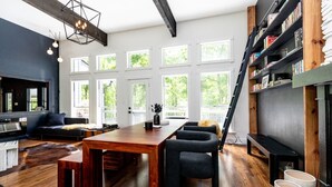A modern living room with a wooden dining table, black sofas, and a bookshelf.