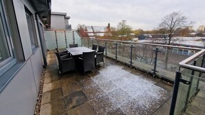 Un balcon avec une table et des chaises, une balustrade en verre et une vue sur les immeubles et les arbres.