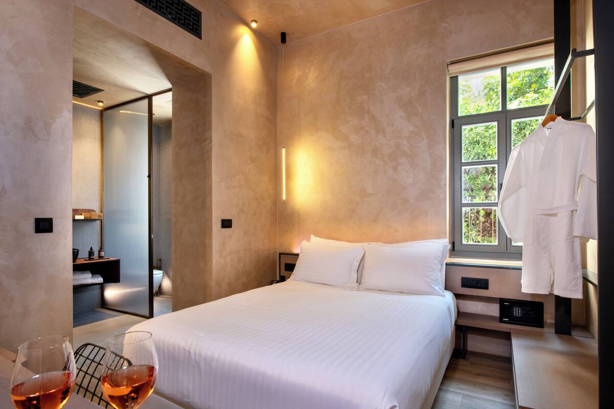 Ein modernes Hotelzimmer mit einem großen Bett, einem Nachttisch, einem Kleiderhaken und einem Fenster mit Blick auf Grünflächen.