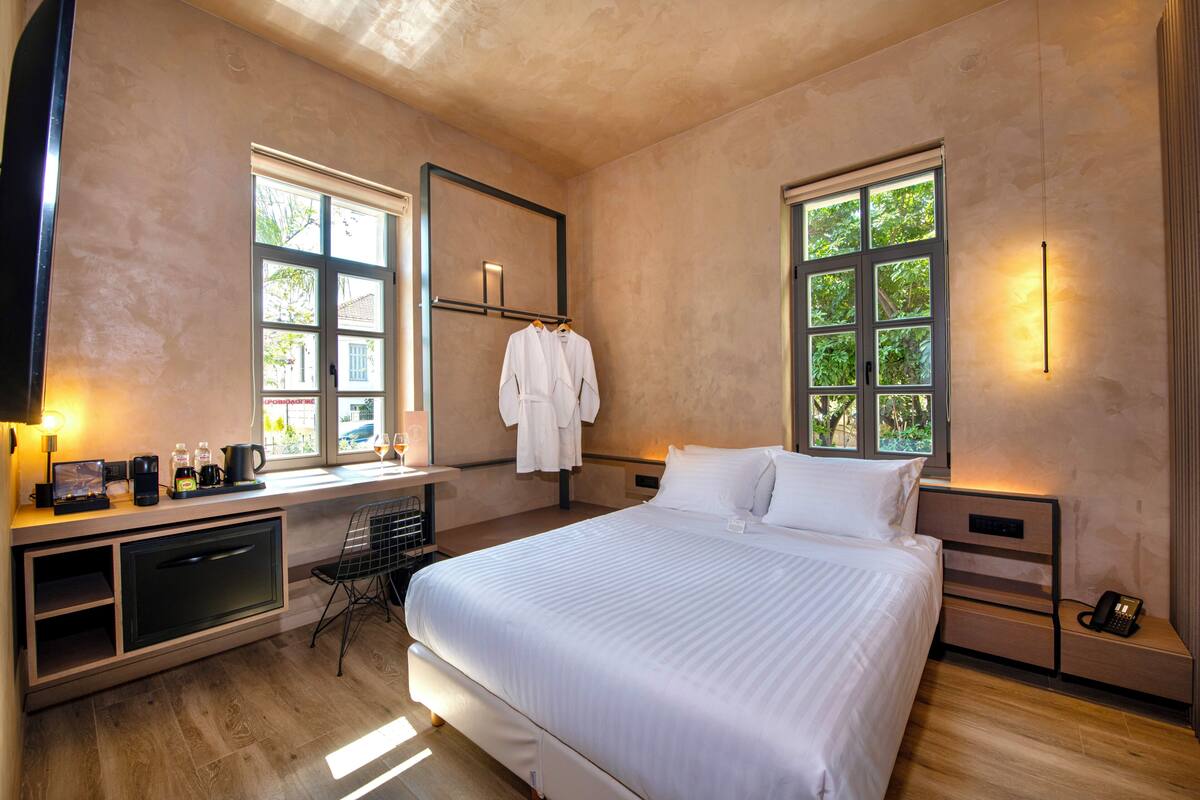 Ein modernes Hotelzimmer mit einem großen Bett, einem Schreibtisch mit Stuhl, einem Kleiderschrank und zwei Fenstern, die für natürliches Licht sorgen.