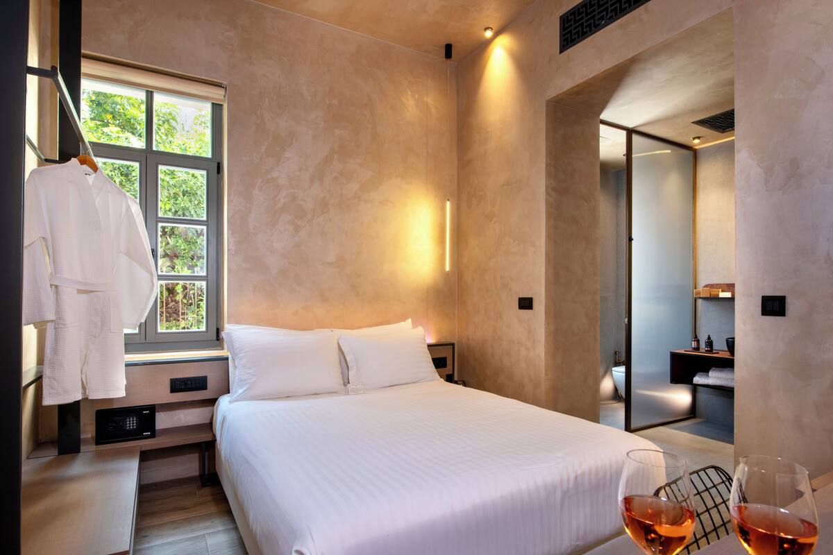 Ein modernes Hotelzimmer mit einem großen Bett, einem Fenster mit Blick auf Grünflächen, einem Bademantel, der am Fenster hängt, und einem Glas Wein auf dem Nachttisch.