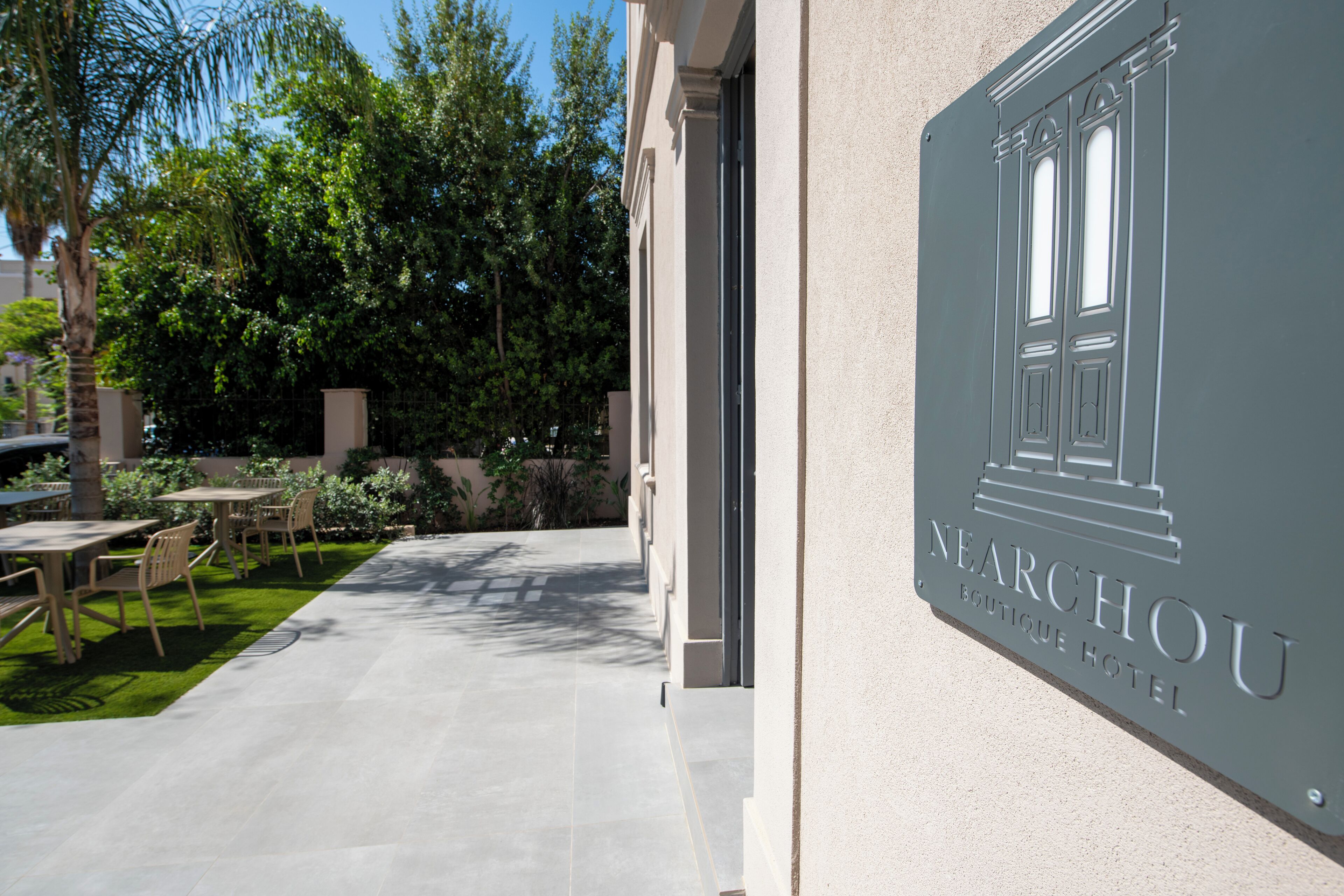 A hotel entrance with a sign reading "NEARCHOU BOUTIQUE HOTEL" and a paved outdoor area with tables and chairs.