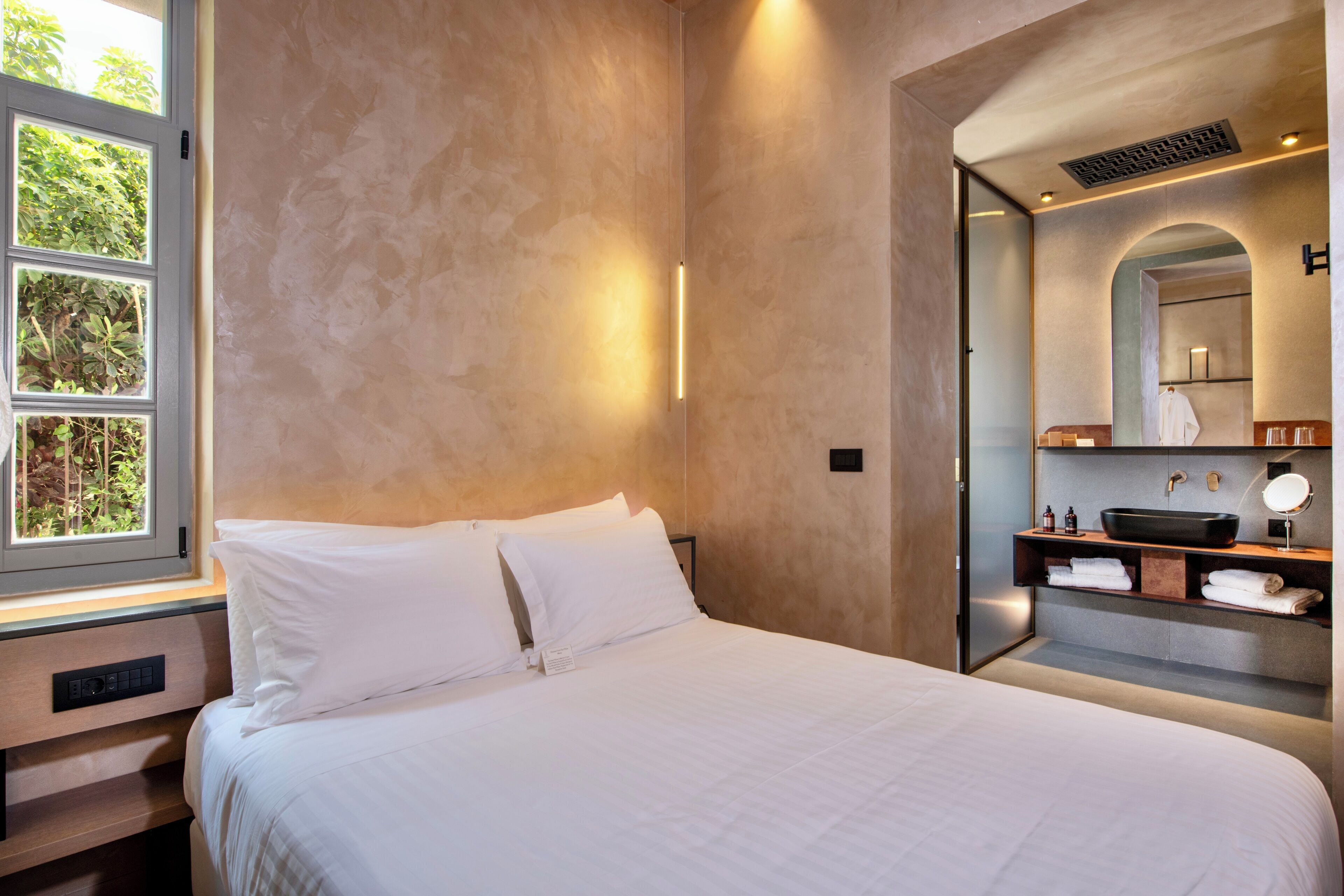 A hotel room with a large bed, a built-in desk, a vanity with a mirror, and a window with a view of greenery.