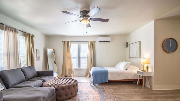 A modern hotel room with a sofa, ottoman, bed, and a small bedside table with a lamp. - The Loft - 1BR/1BA - Close to TTU (Lubbock)