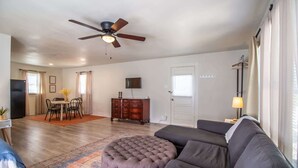A living room with a sofa, ottoman, ceiling fan, dining area, and a TV mounted on the wall. - The Loft - 1BR/1BA - Close to TTU (Lubbock)