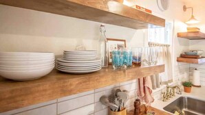 Una cocina con estantes de madera que sostienen platos, vasos y una botella. Hay utensilios en un organizador y un lavamanos con un grifo.