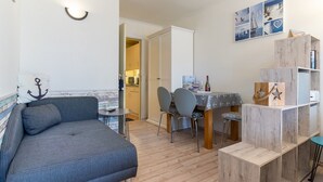 A cozy living area with a sofa, dining table, chairs, and a wooden bookshelf. - Ferienwohnung in St. Peter-ording (St. Peter-Ording)