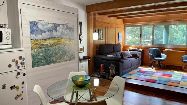 A modern living room with a glass dining table, white chairs, a black sofa, and a colorful quilt. - Beautiful studio, fully equipped.   (La Molina)