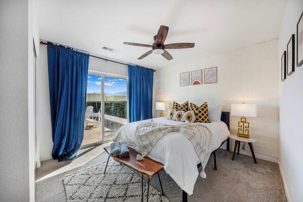 A bedroom with a bed, a ceiling fan, a nightstand with a lamp, and a view of the outdoors.