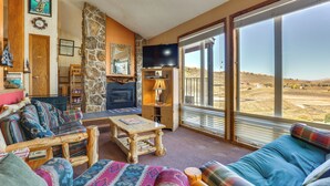 A cozy living room with a stone fireplace, a flat-screen TV, and large windows offering a view of the countryside. - Walk to Ski Lift & Hiking: Granby Ranch Condo! (Granby)