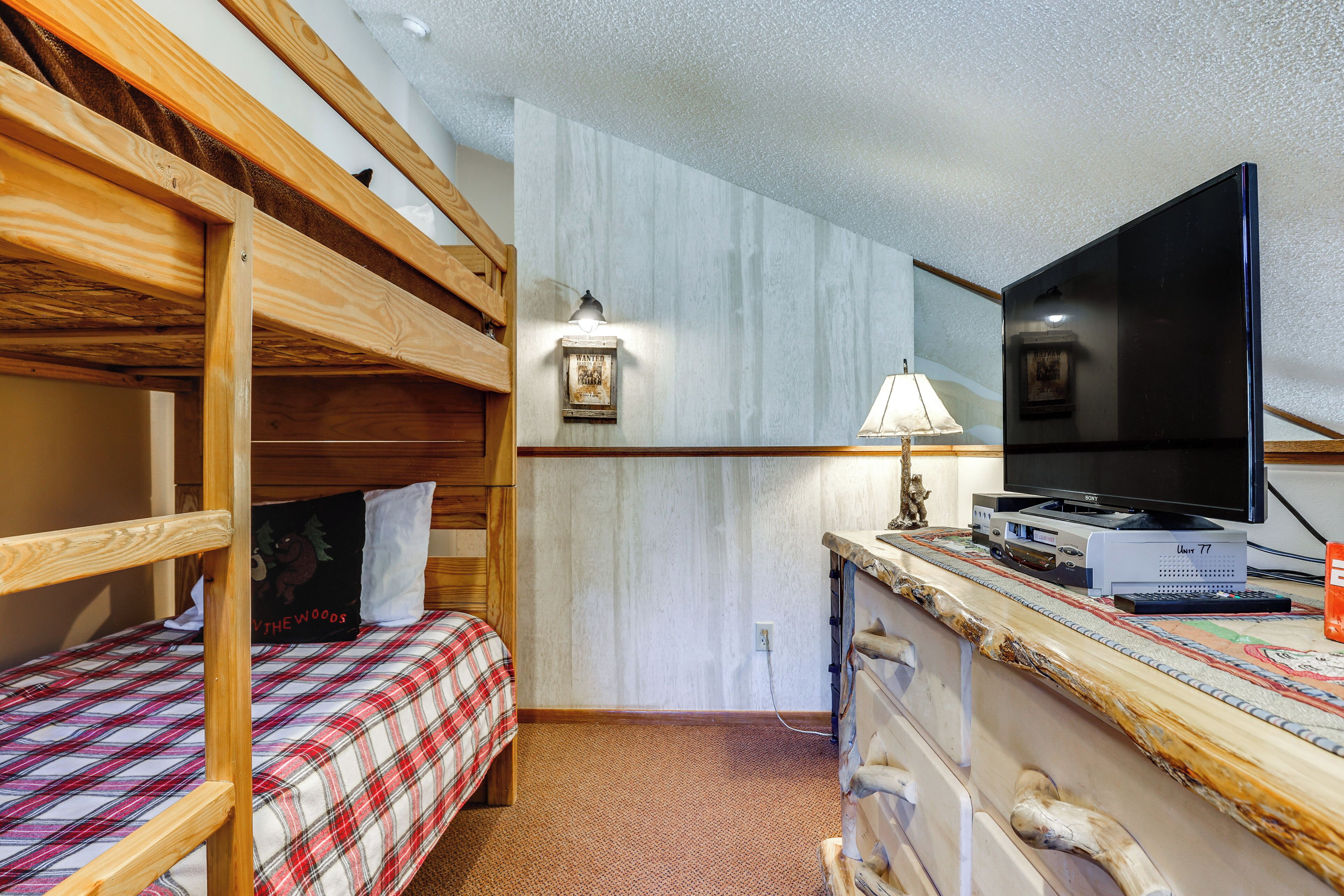 A bunk bed with a plaid bedspread, a desk with a lamp, a flat-screen TV, and a picture frame on the wall.