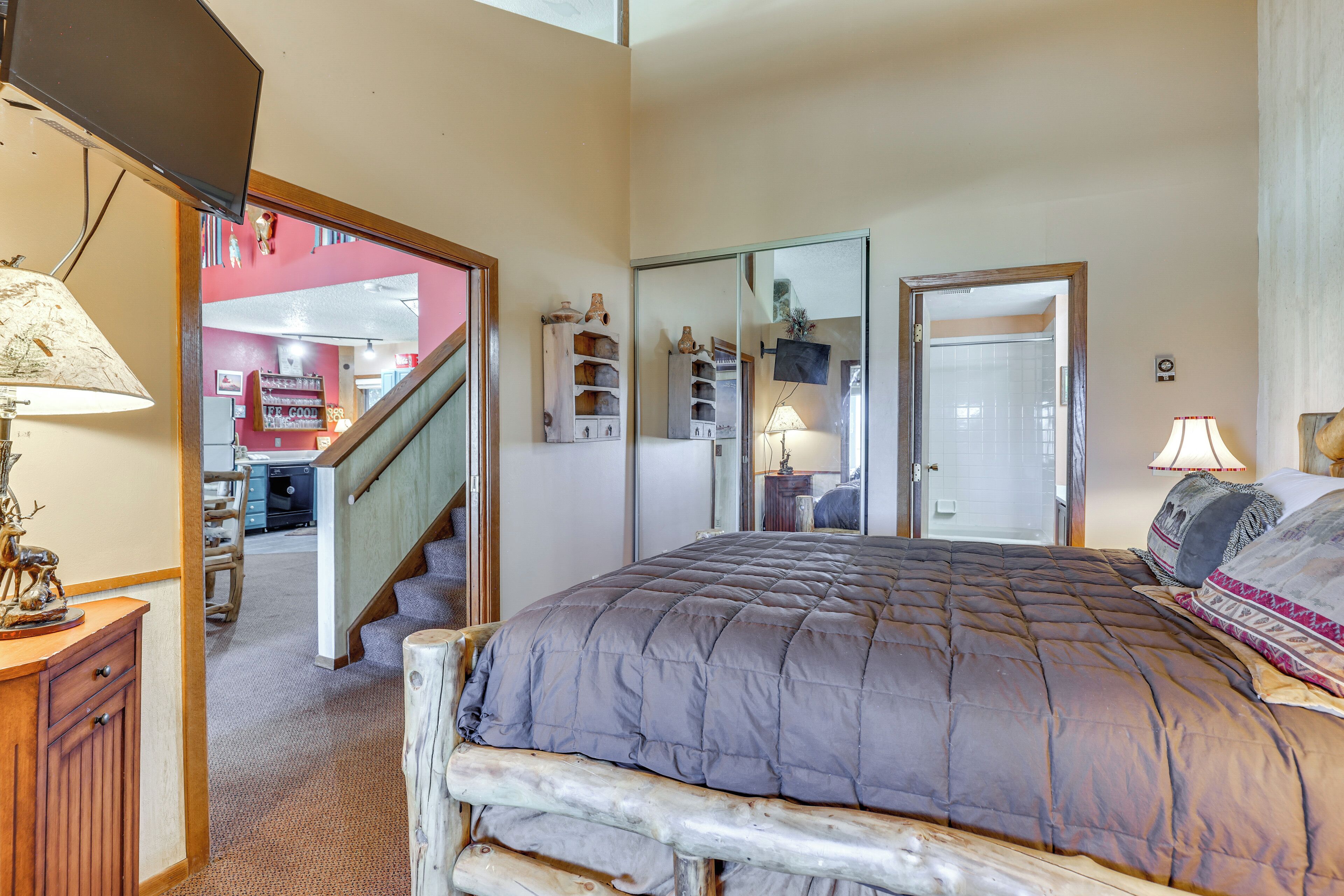 A bedroom with a large bed, a wooden headboard, a desk, and a chair. There is a mirror on the wall and a staircase leading to another room.