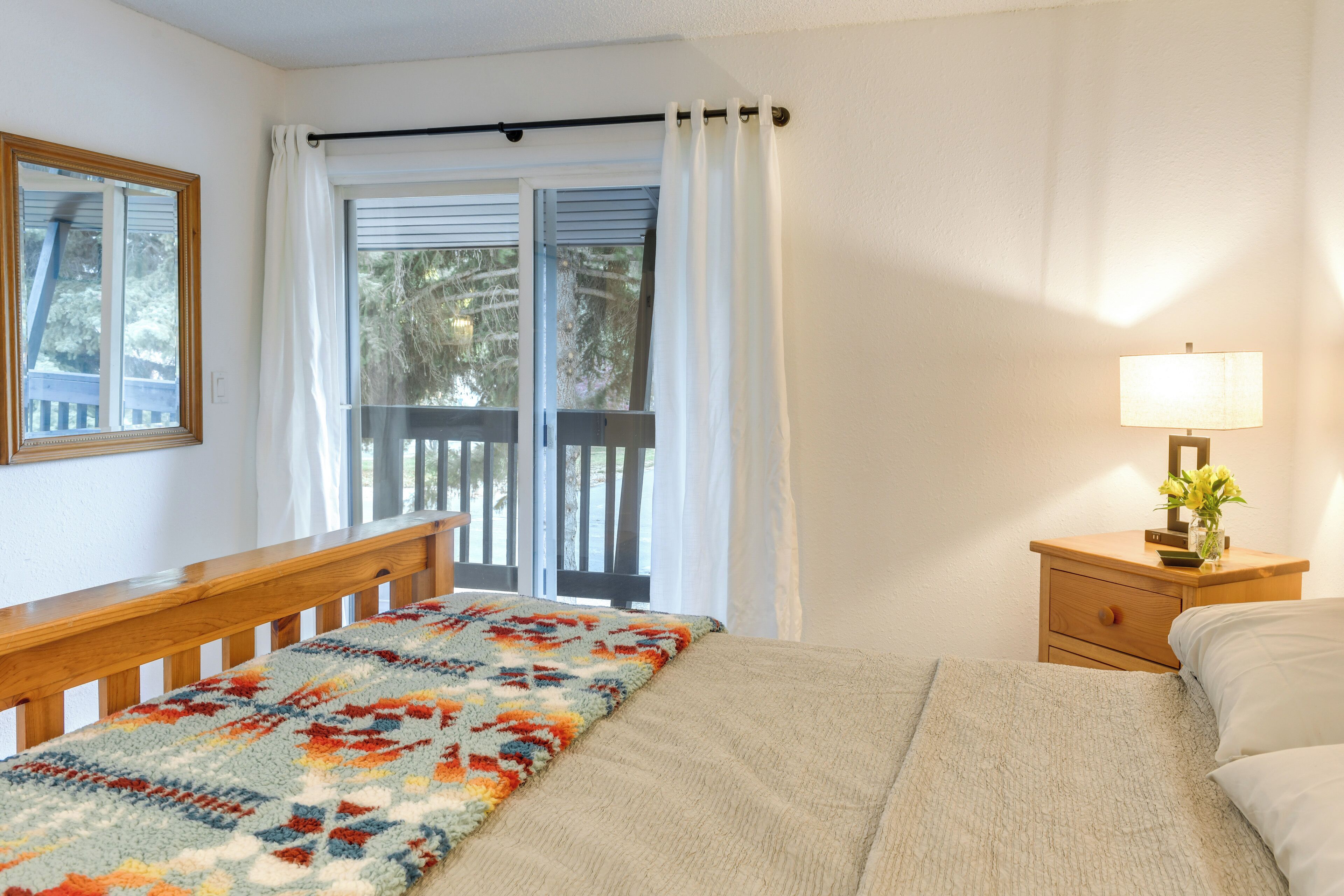 Une chambre à coucher avec un lit, une table de chevet avec une lampe, une fenêtre avec des rideaux et une balustrade en bois à l’extérieur.