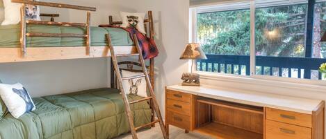 A bunk bed with a ladder, a desk with drawers, a lamp, and a window with a view of trees.