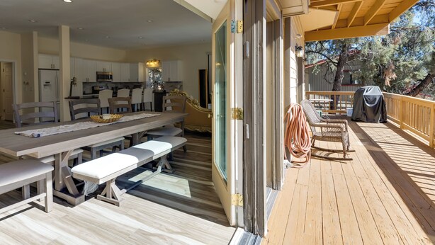 A spacious indoor dining area with a long table and benches, a kitchen in the background, and a wooden deck with outdoor seating. - Game Room & Deck: Modern Frazier Park Home! (Pine Mountain Club)