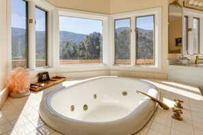 Eine große, ovale Badewanne in einer Ecke, mit drei Fenstern, die Blick auf einen bewaldeten Hügel bieten.