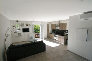 A modern kitchen with a flat-screen TV, a dining area with a black sofa, and a view of greenery outside.