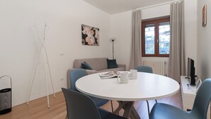 Egy modern nappali, melyben egy kerek, fehér asztal, kék székek, egy bézs színű kanapé és egy falfestmény található.