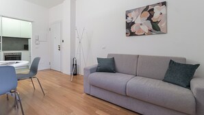 Egy modern nappali, szürke színű kanapéval, étkezőasztallal és hozzáillő székekkel, valamint fehér szekrényekkel berendezett konyhasarokkal.