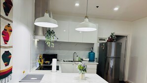 Eine moderne Küche mit weißen Schränken, einer Marmorarbeitsplatte, zwei Pendelleuchten, einem Edelstahlkühlschrank und einer kleinen Kücheninsel mit einer Blumenvase.