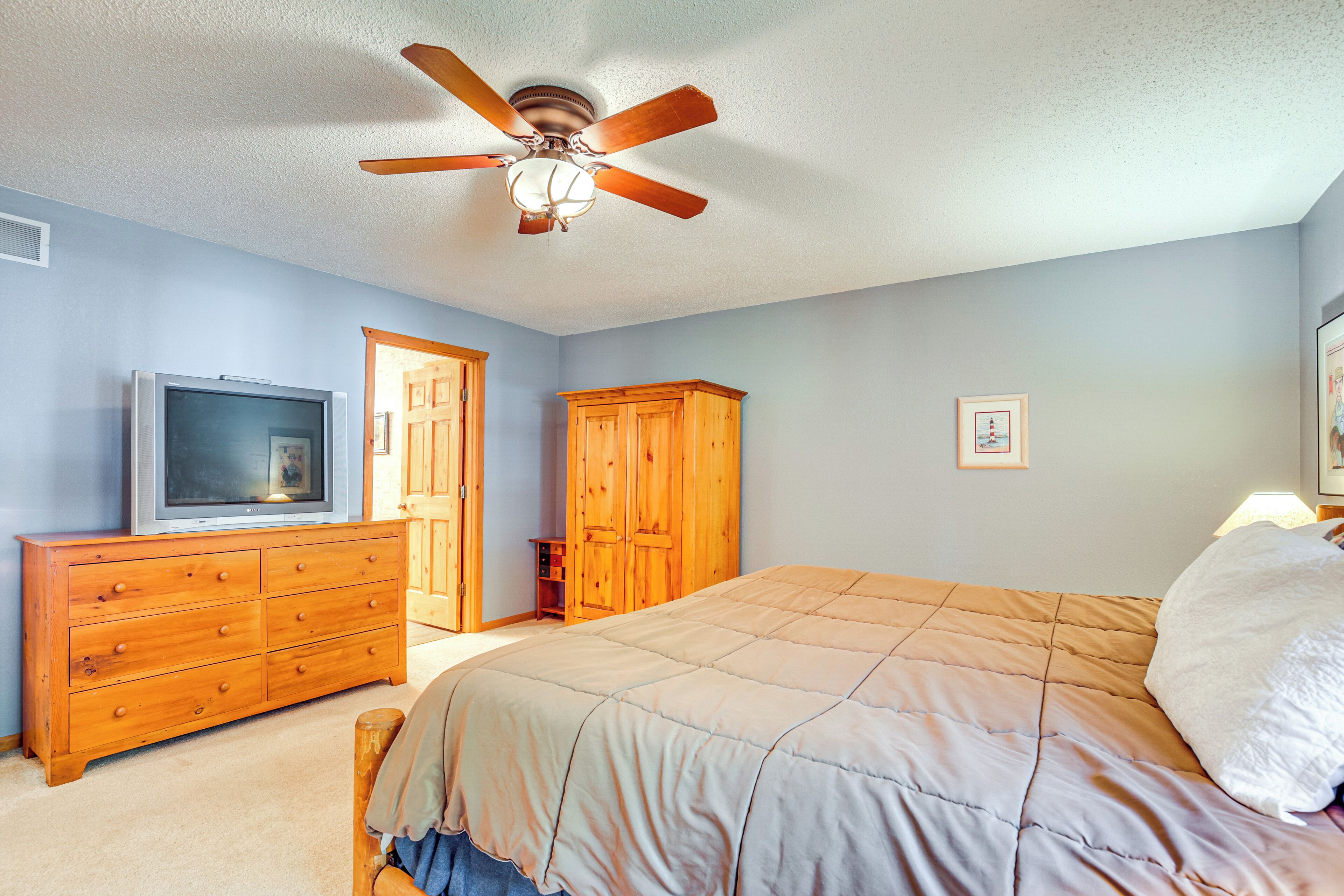 A bedroom with a bed, wooden dresser, television, wardrobe, and ceiling fan.