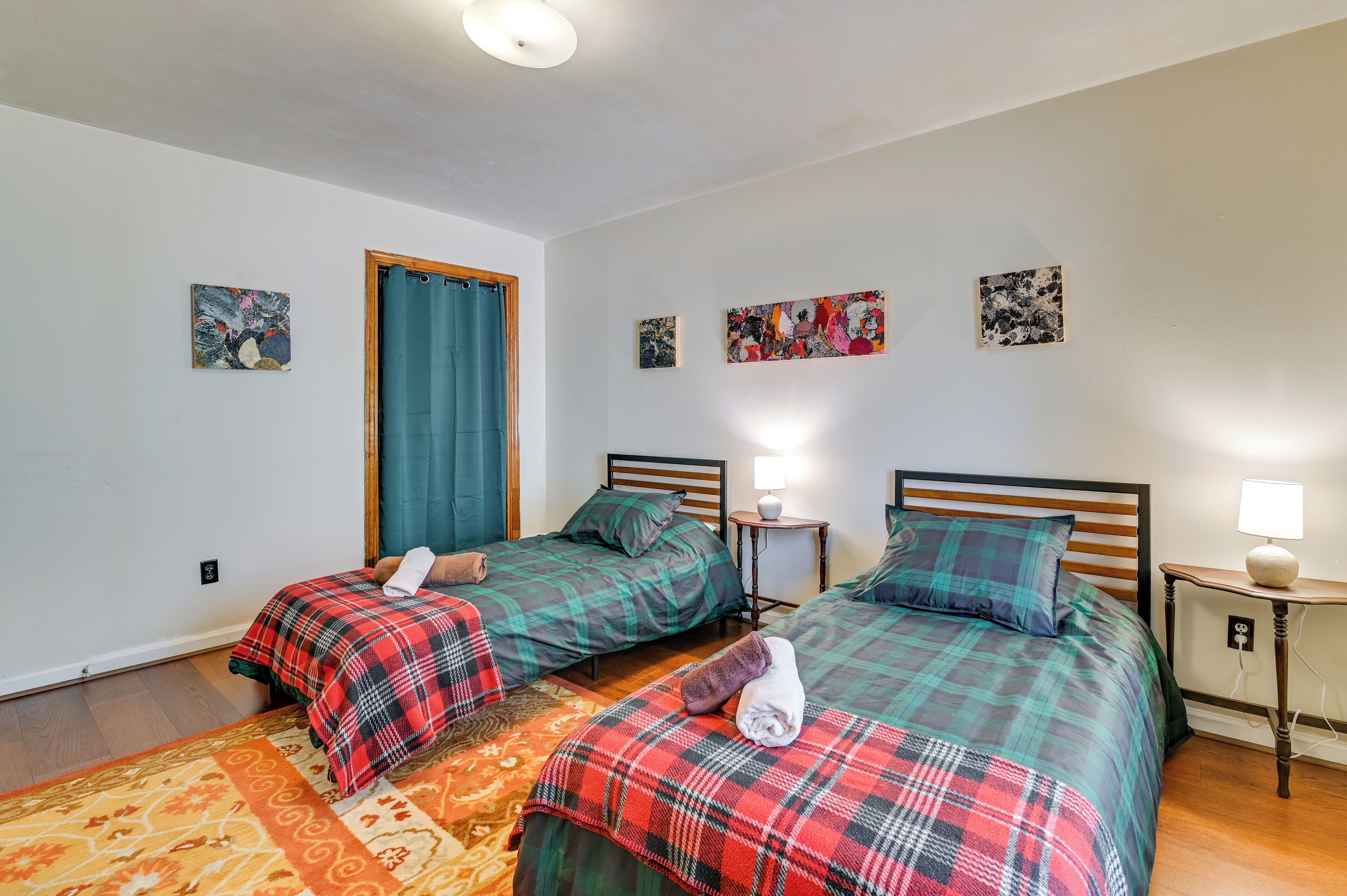 A room with two beds, each with a plaid bedspread, a small bedside table with a lamp, and a wall-mounted light.