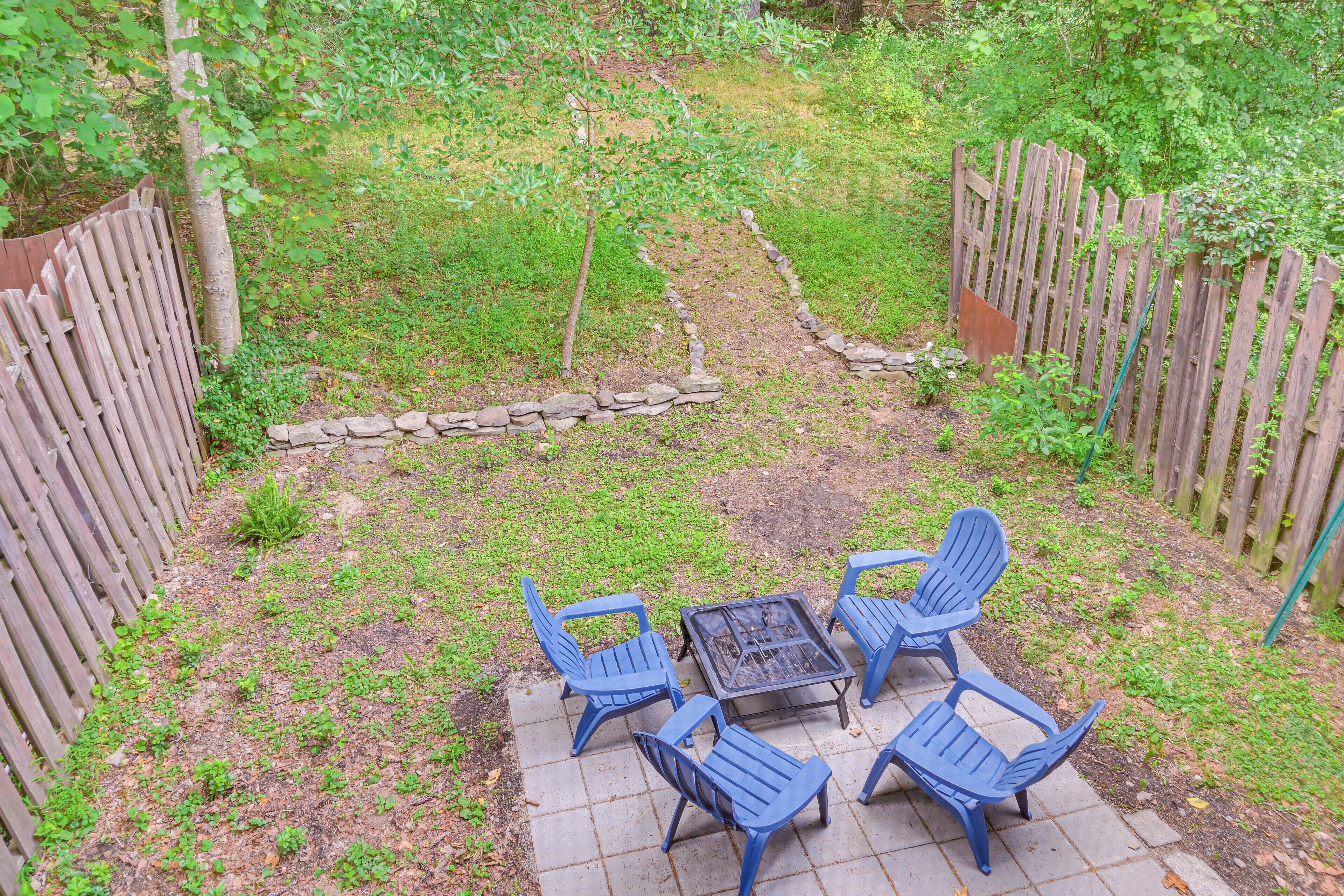 A patio with a fire pit and four blue chairs, surrounded by a wooden fence and a stone border.