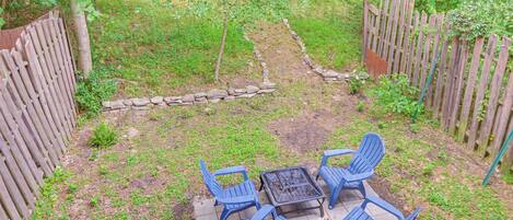 A patio with a fire pit and four blue chairs, surrounded by a wooden fence and a stone border.