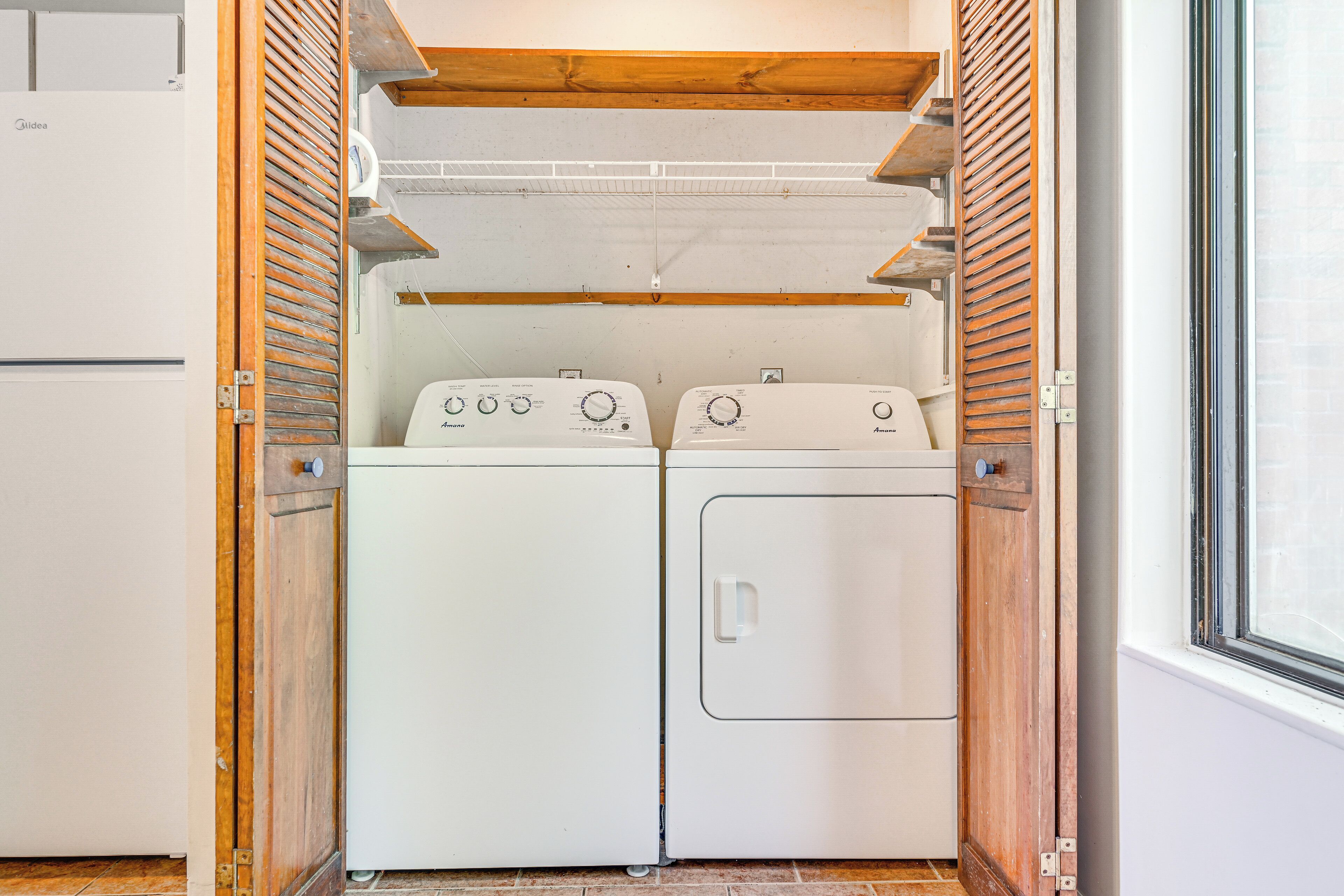 A laundry room with two white washing machines, wooden slatted doors, and a window on the right.