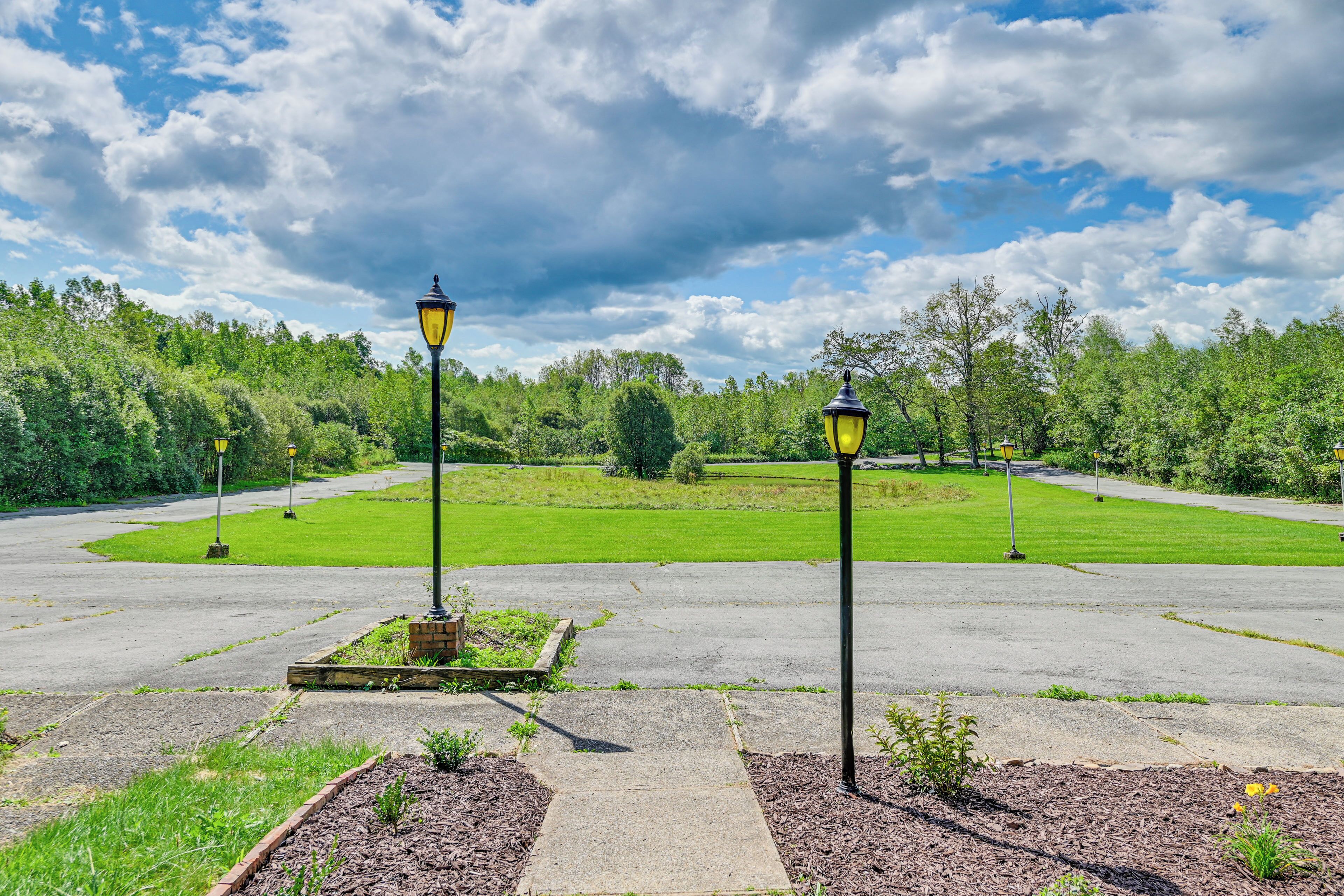 Un espace de stationnement dallé, avec deux lampadaires et, au fond, un champ d’herbe.