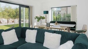 Un salon moderne avec un canapé vert, des coussins blancs et moelleux, une table à manger avec des chaises, un vase de fleurs et une vue sur un jardin et un immeuble, le tout visible à travers de grandes fenêtres.