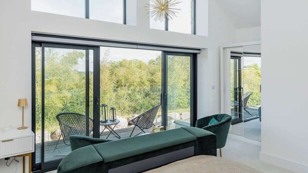 Une chambre moderne dotée de grandes fenêtres, d’un lit, d’un bureau et d’un coin salon avec des fauteuils.