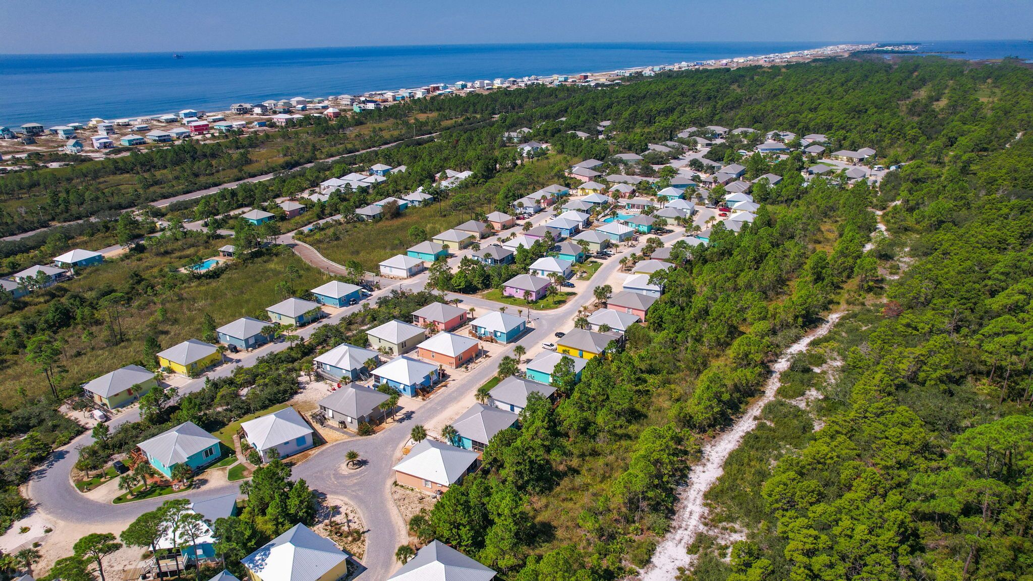 Vista aérea de un área residencial con casas alineadas en filas, rodeada de abundante vegetación y una costa en el fondo.