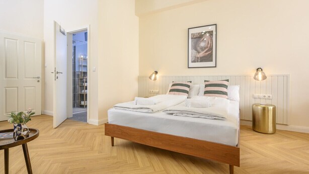 A bedroom with a wooden bed, bedside lamps, a framed picture on the wall, and a small round table with a plant. - Palais Palffy Hotel Suite in Central Vienna (Vienna)