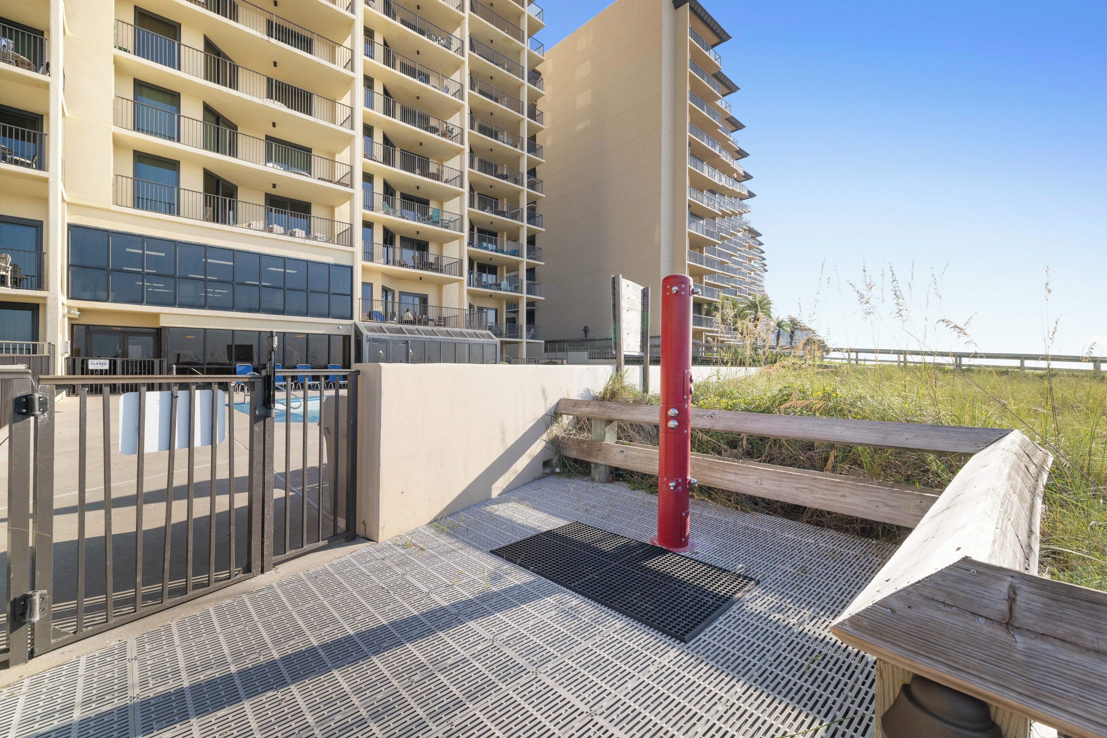 Une entrée sécurisée, avec portail, donnant sur un immeuble de grande hauteur doté de nombreux balcons, d’un poteau rouge et d’une terrasse en bois.