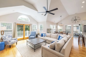 A spacious living room with a high ceiling, a ceiling fan, a large window, a dining area, and a kitchen. - Oceanside Duck Retreat w Pool, Spa, Mins to Beach! (Duck)