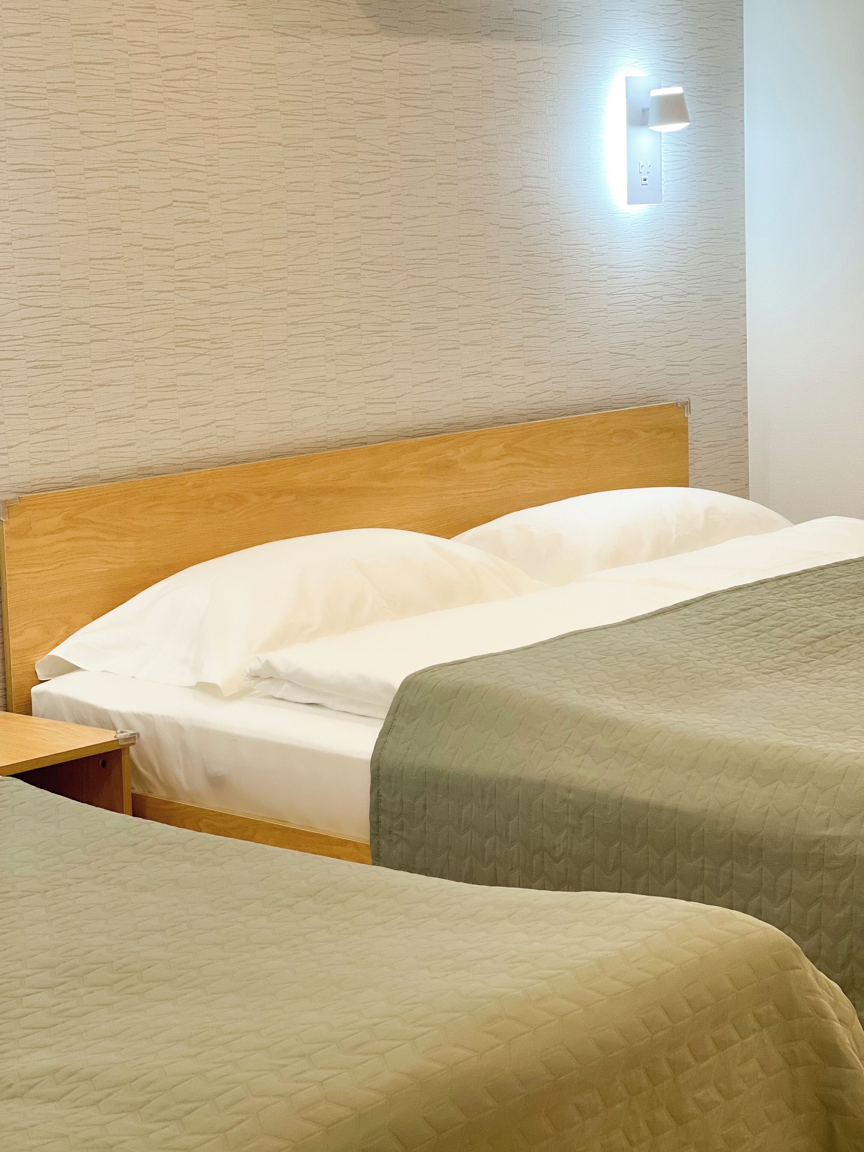 Een hotelkamer met een houten hoofdeinde, twee bedden met groen en witte beddengoed, een nachtkastje en een wandlamp.