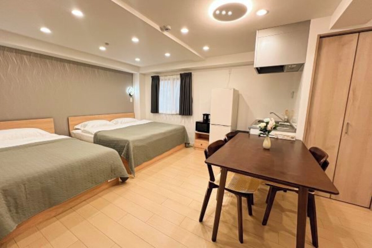 A hotel room with two single beds, a wooden dining table, a small kitchenette, and a wardrobe.