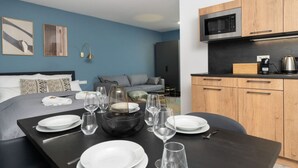 Een moderne hotelkamer met een net gedekte eettafel, een bed met gestreepte kussens, een blauwe wand en een kitchenette met houten kastjes.