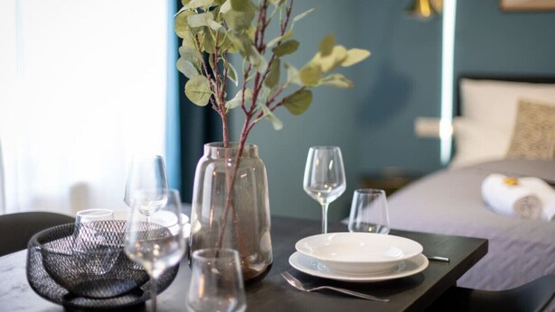 Een net gedekte eettafel met een vaas groene bladeren, wijnglazen en borden, wat duidt op een eetgedeelte in een hotelkamer.
