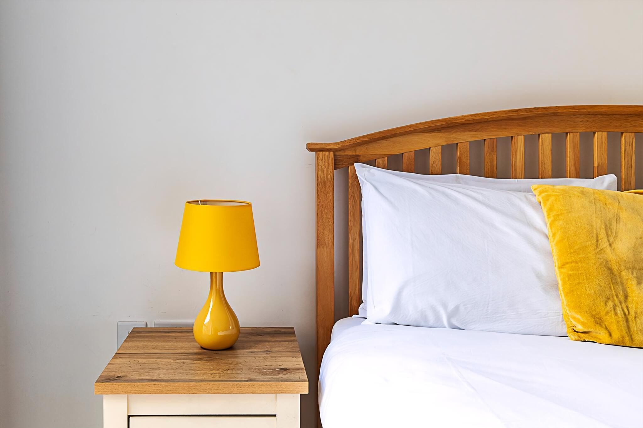A bed with white bedding and a yellow pillow, a wooden nightstand with a yellow lamp, and a wooden headboard.